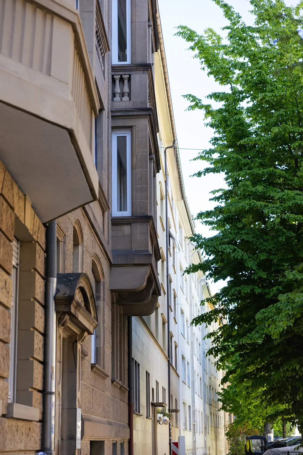 Altbaufassade mit vorspringendem Erker, dahinter eine baumbestandene Straße.